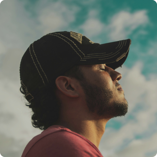 A man in a black cap and pink shirt stands outdoors with his eyes closed, facing upward against a cloudy sky.