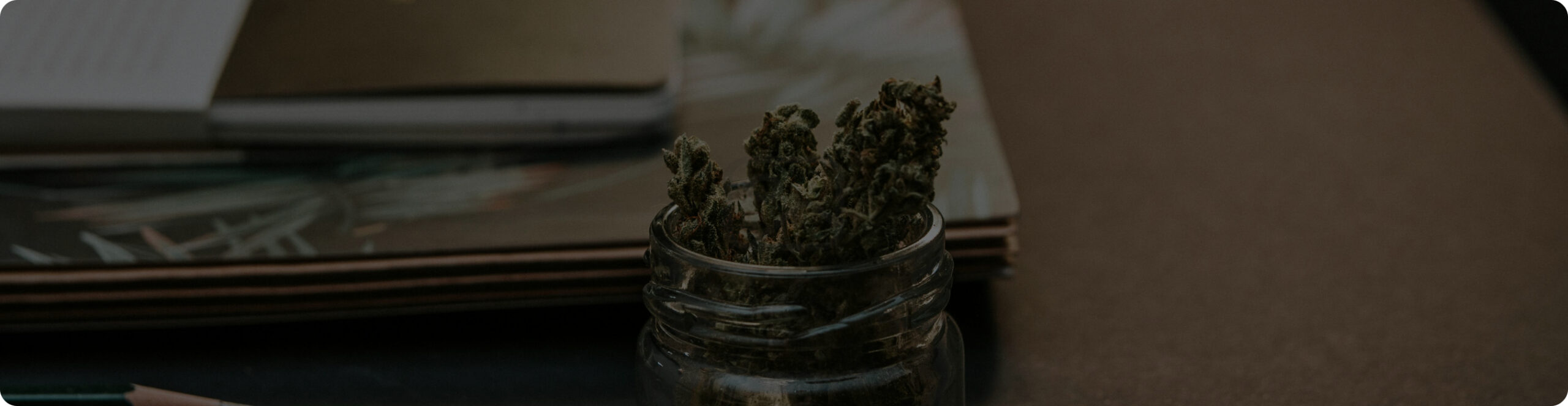 A small glass jar containing dried herb sits on a table next to stacked books and a notebook.
