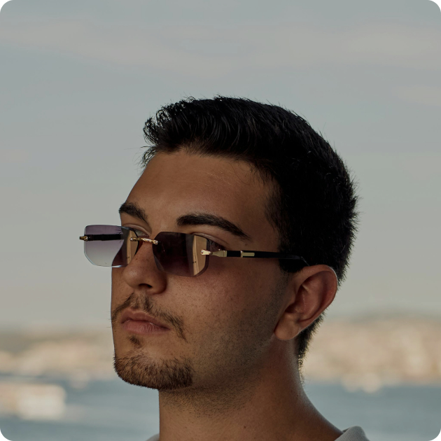 A man with short dark hair and a trimmed goatee wears rectangular sunglasses, looking into the distance with a blurred outdoor background.