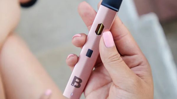 A hand holding a pink vape pen with a "B." on the bottom, showing manicured nails with light pink polish.