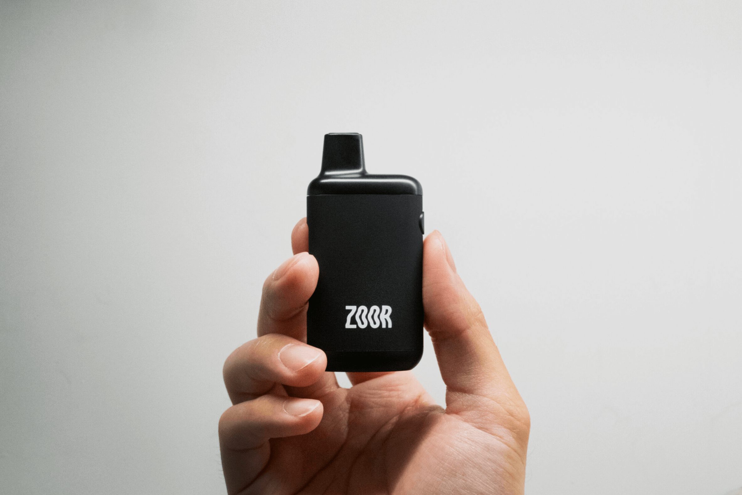 A hand holds a black vaping device labeled "ZOOR" against a plain white background.